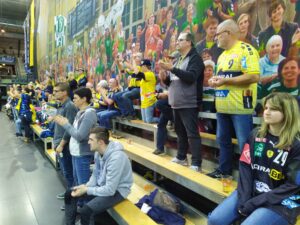 Fans der Rhein-Neckar Löwen auf der kleinen Tribüne mit Holzbänken in der Arena Leipzig, vor einem riesigen Transparent mit jubelnden Fans des SC DHfK Leipzig.